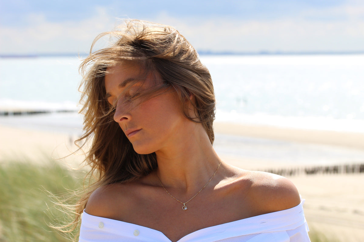 Woman at the beach, eyes closed, wearing the small Heart to Heart breast milk pendant in yellow gold