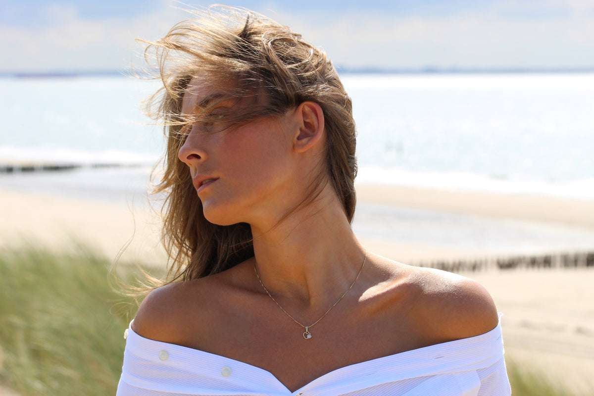 Woman at the beach, eyes closed, wearing the small Heart to Heart breast milk pendant in yellow gold
