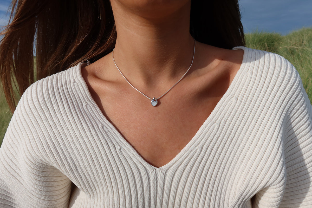 Close-up of the silver Amore heart breast milk pendant on a snake chain with a white V-neck sweater