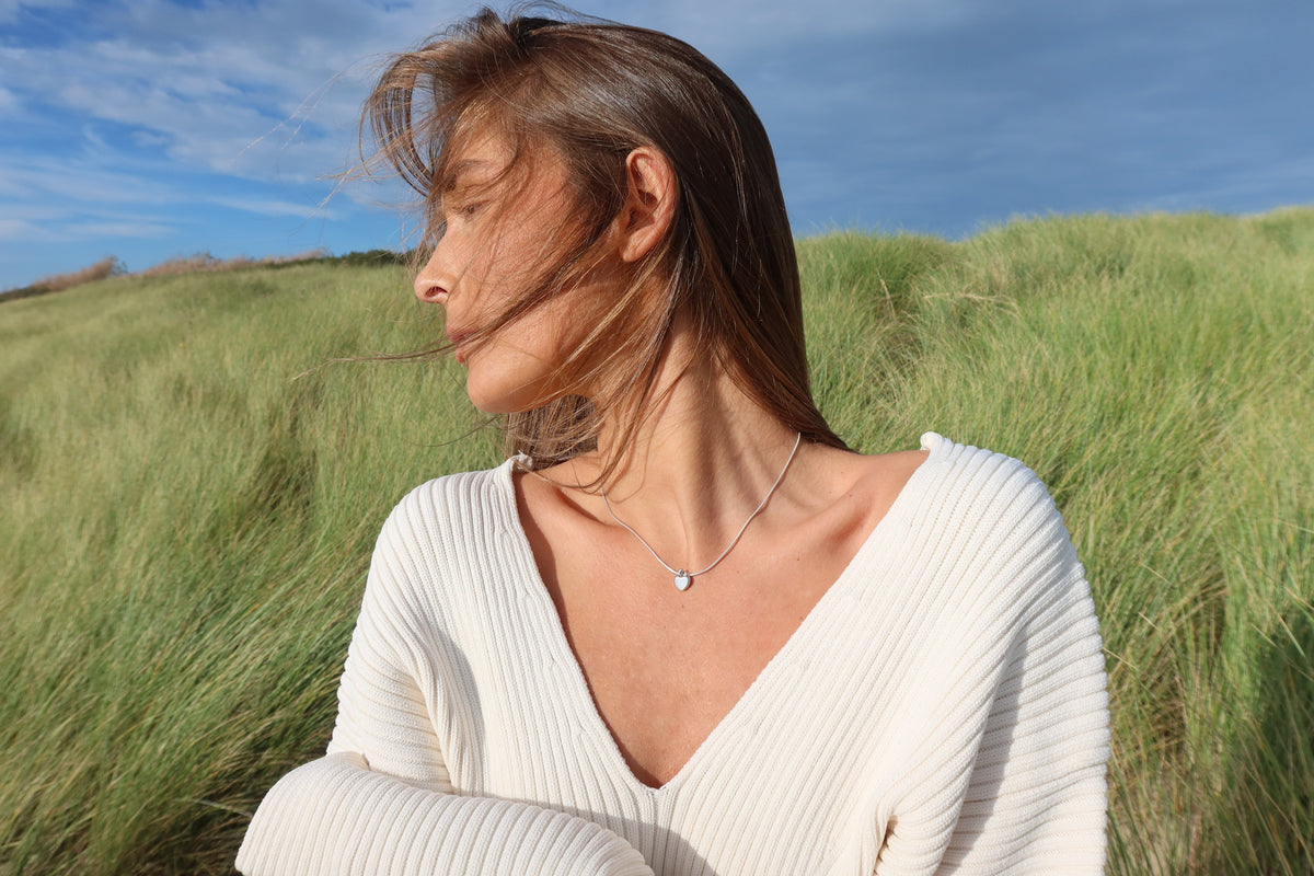 Woman in a white knit sweater in the dunes wearing the silver Amore heart breast milk pendant
