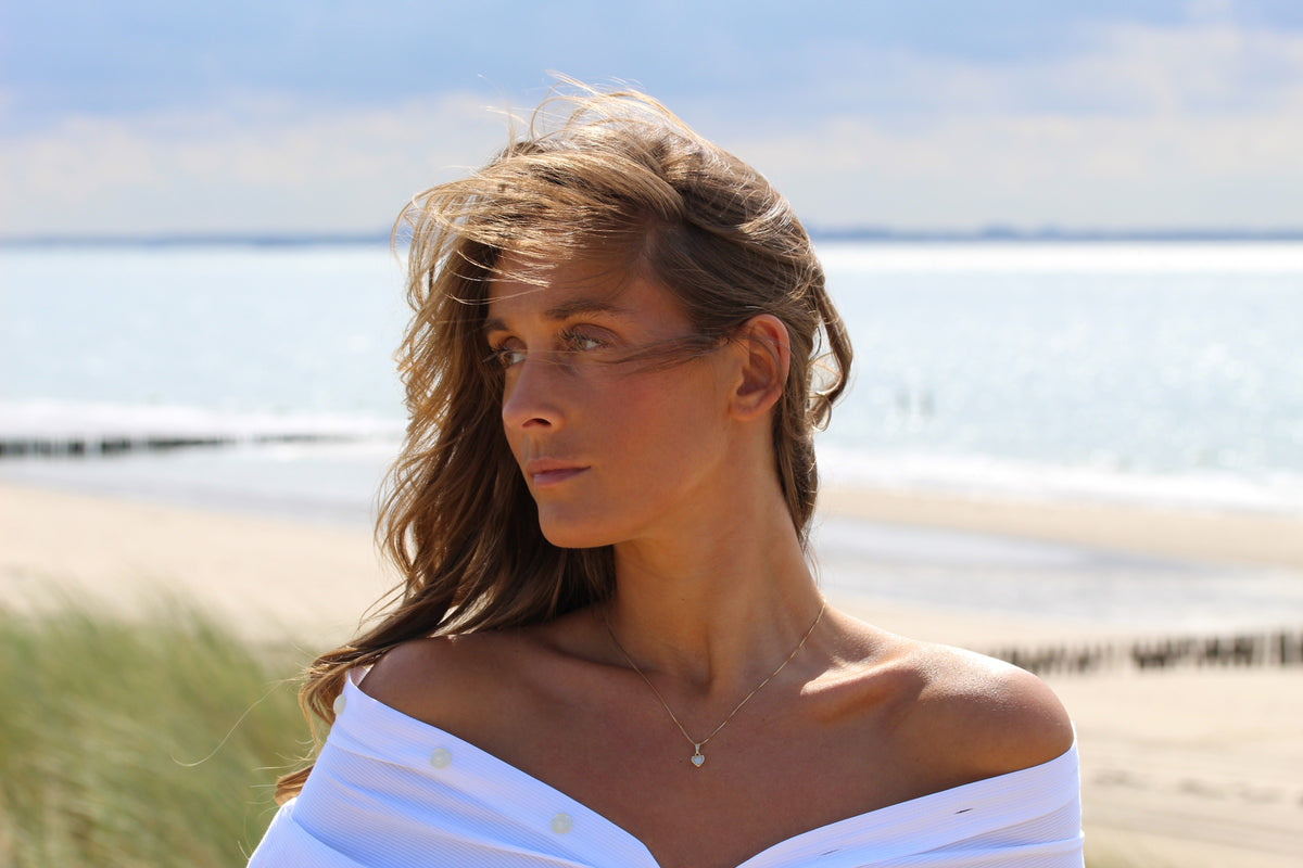 Woman at the beach in an off-shoulder white shirt wearing the small Cara heart breast milk pendant