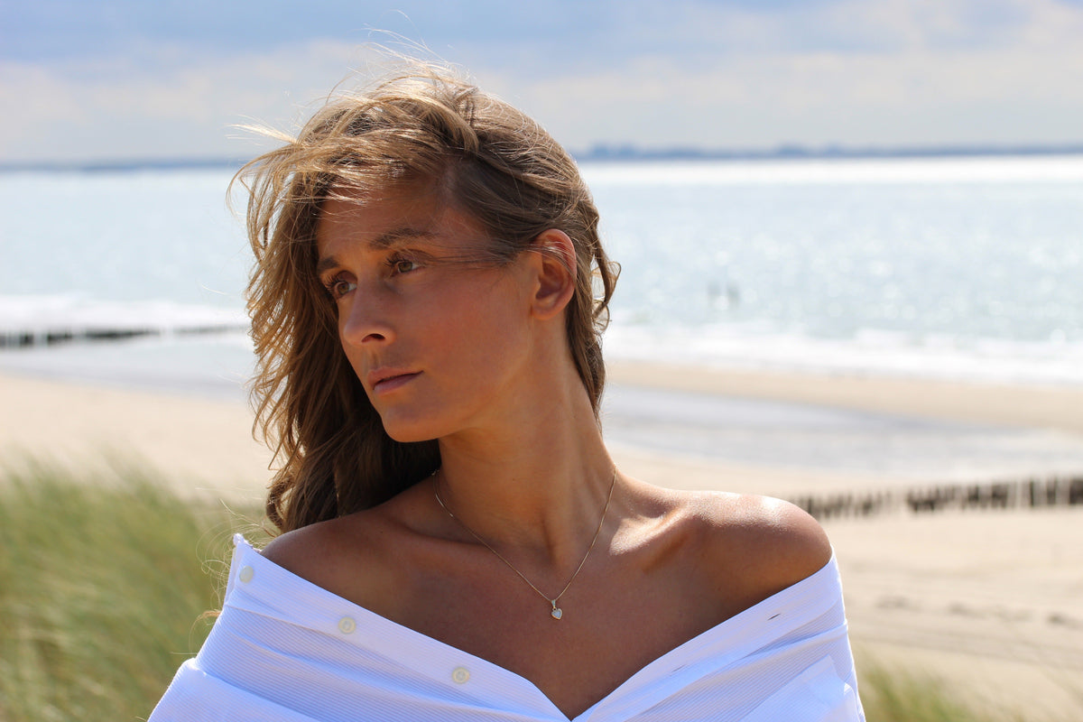 Woman by the sea with windswept hair wearing the Cara heart breast milk pendant on a fine chain