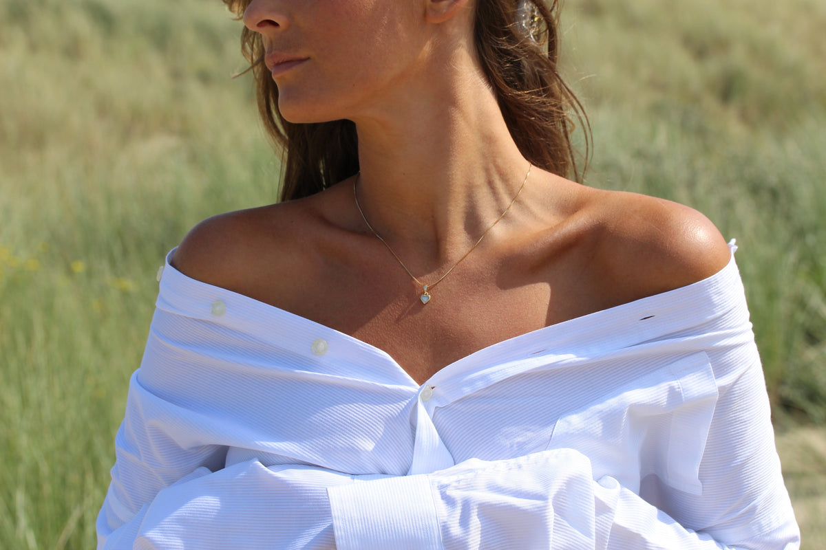 Woman in an off-shoulder white shirt in the dunes wearing the Cara heart breast milk pendant