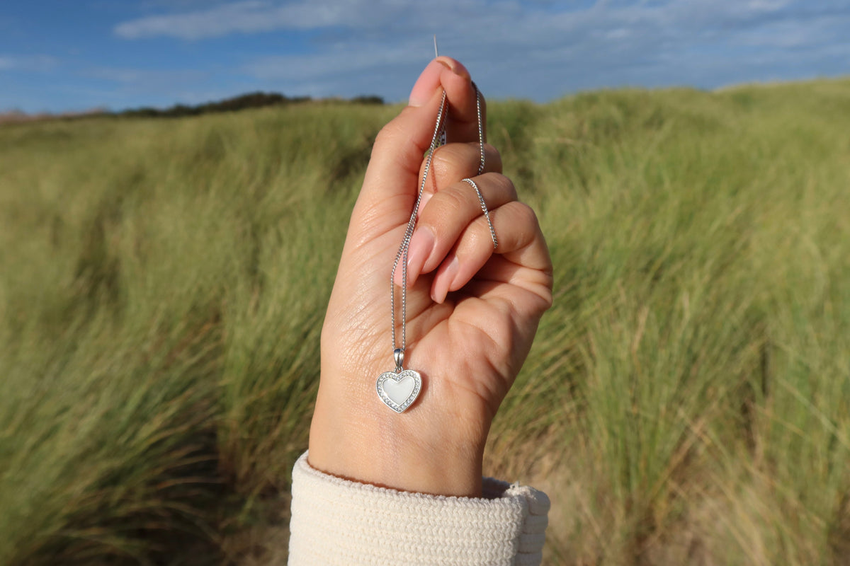 Hand holding a silver-colored Demi halo heart breast milk pendant on a chain in the dunes