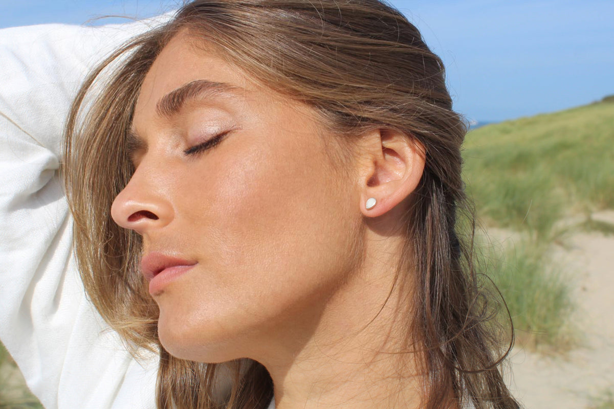 Woman with eyes closed and arm raised on a beach, wearing a silver Drop breast milk stud earring