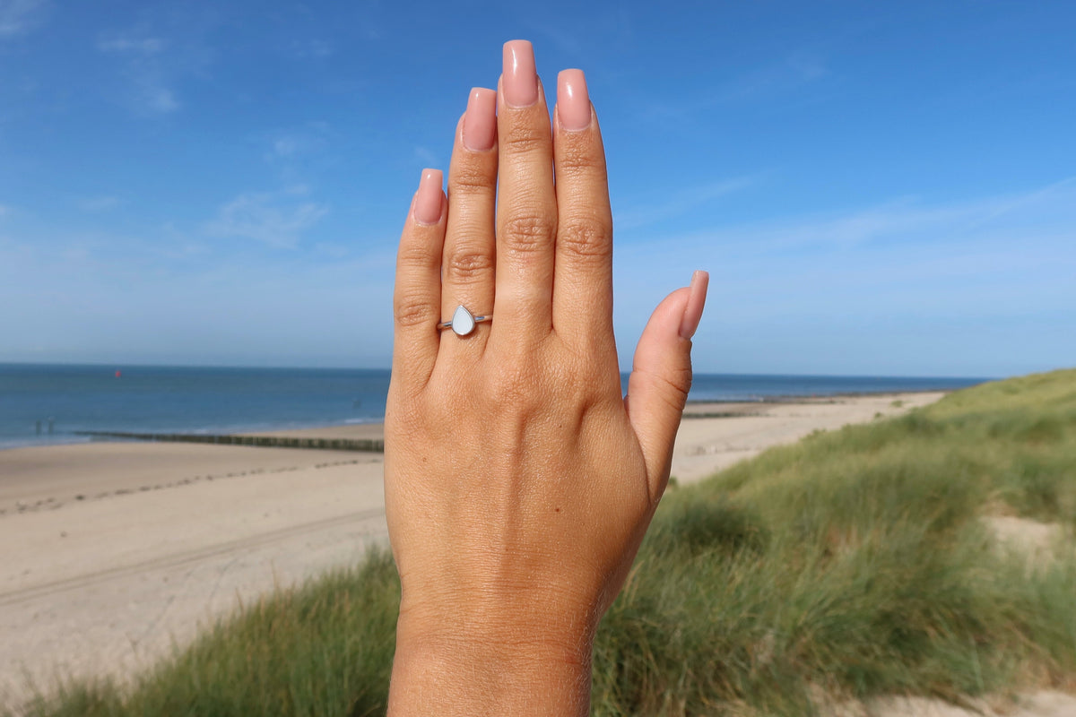Back of a hand raised against the beach and dunes, wearing the silver teardrop Droplet breast milk ring
