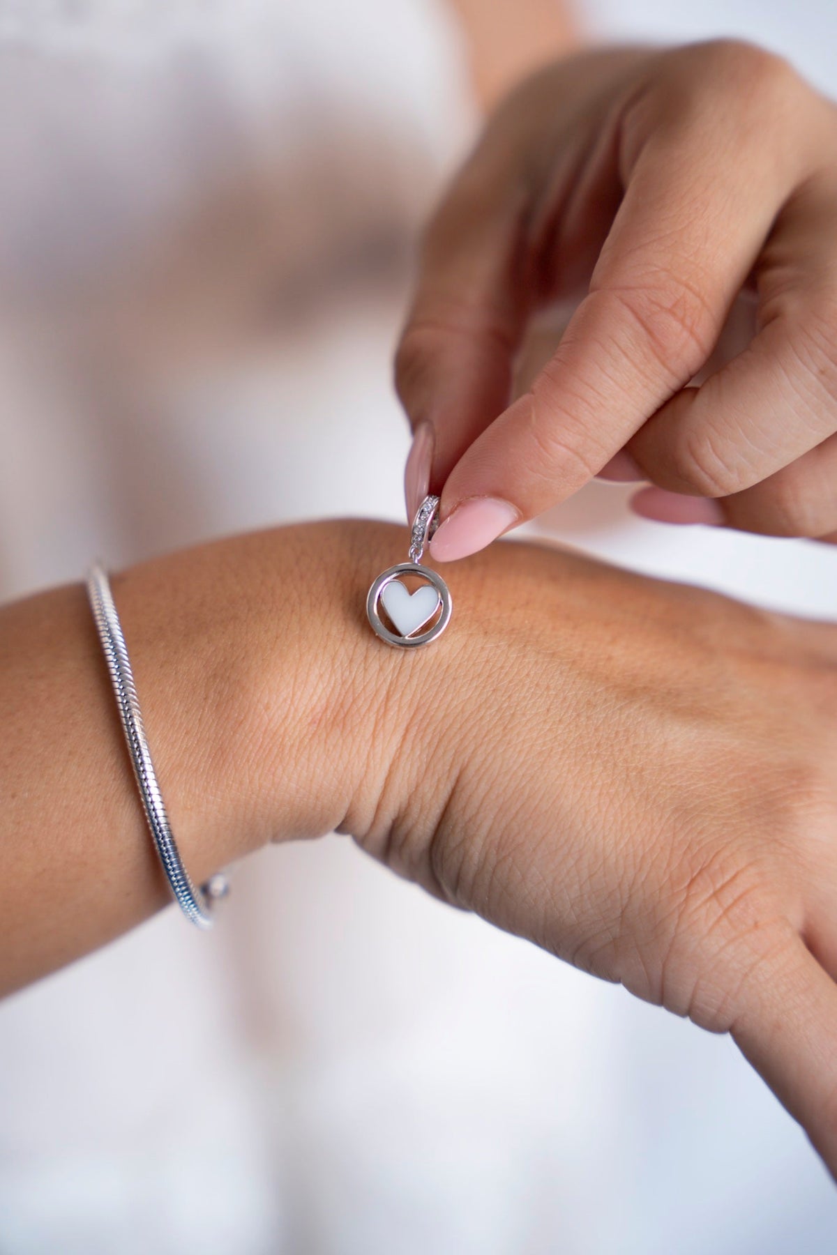Eline breast milk charm in solid white gold with a heart of breast milk in a round frame, next to a bracelet.