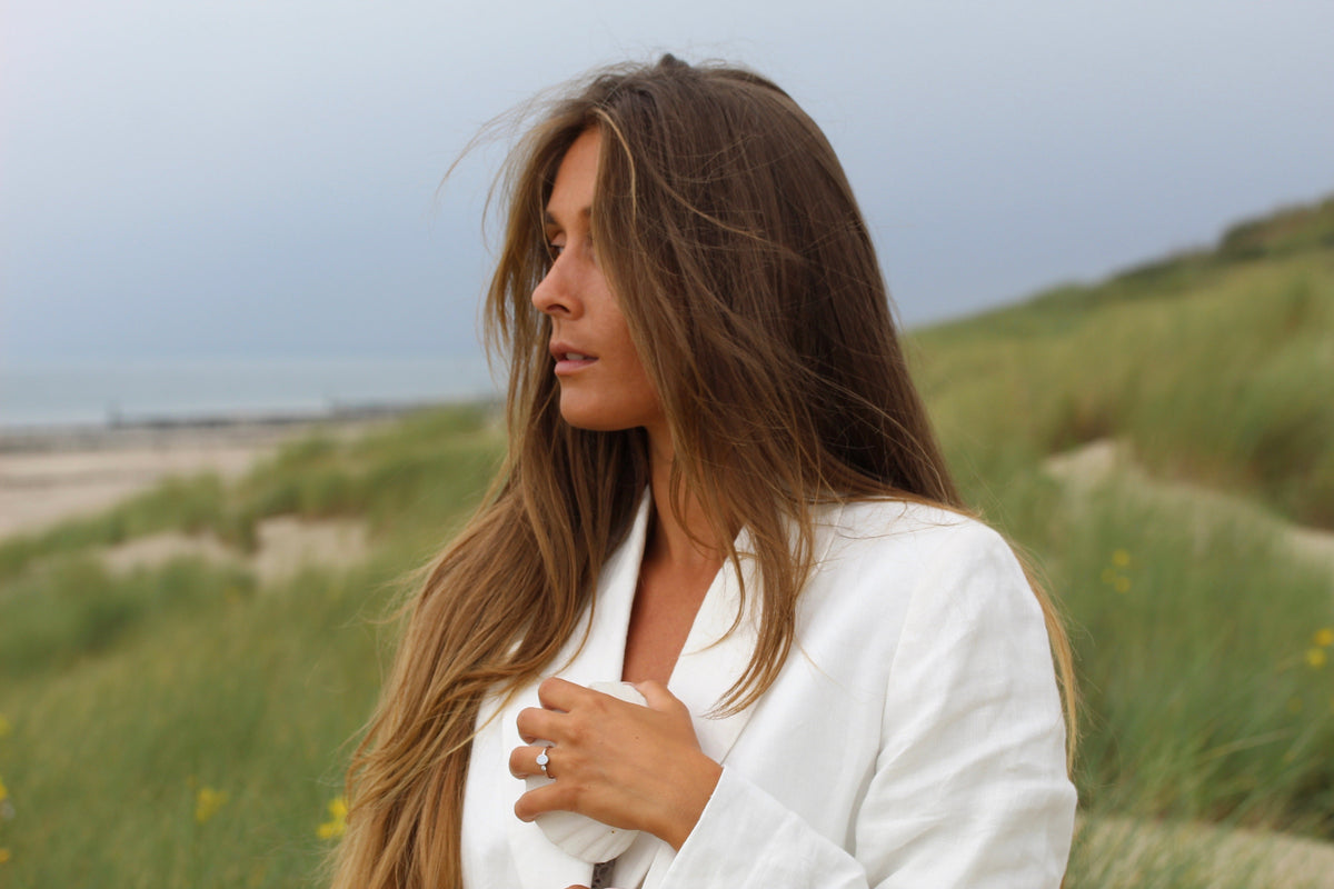 Woman in a white blazer in the dunes holding a shell, wearing the silver Elisa breast milk ring