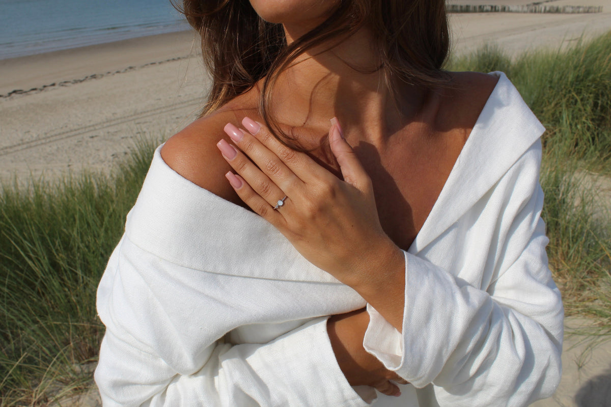 Hand on the chest in a white linen top at the beach, wearing the silver Giulia breast milk ring