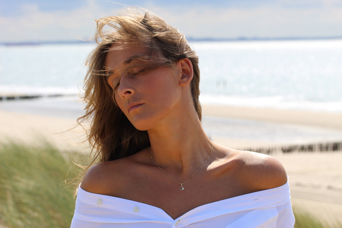 Woman at the beach with windswept hair wearing the Heart to Heart breast milk pendant in yellow gold