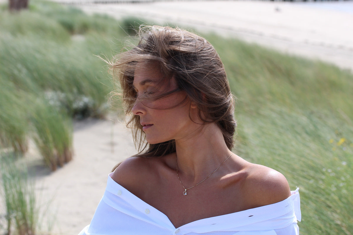 Woman in the dunes wearing the Heart to Heart breast milk pendant in yellow gold, white off-shoulder shirt