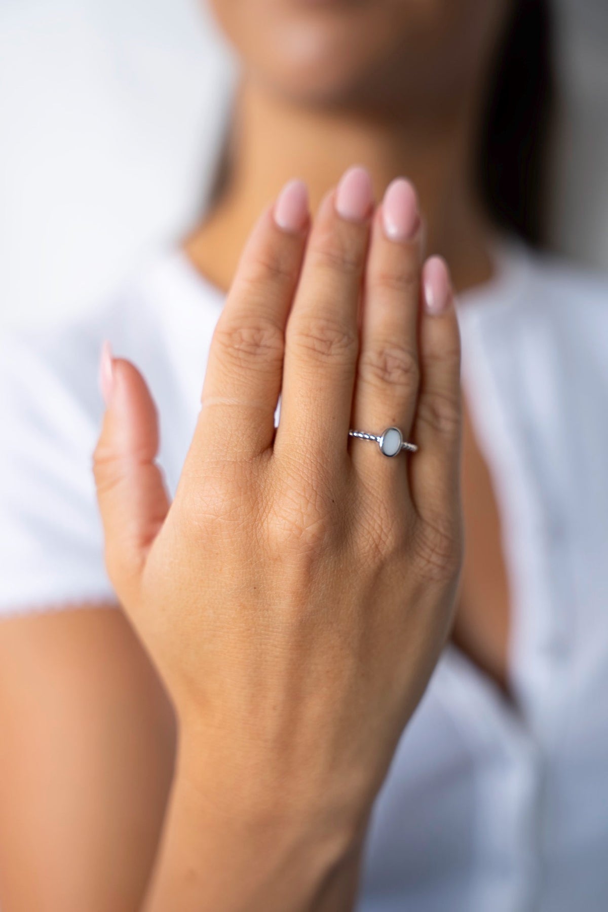 Raised hand wearing the white gold Ilse ring with an oval breast milk stone, against a white shirt.