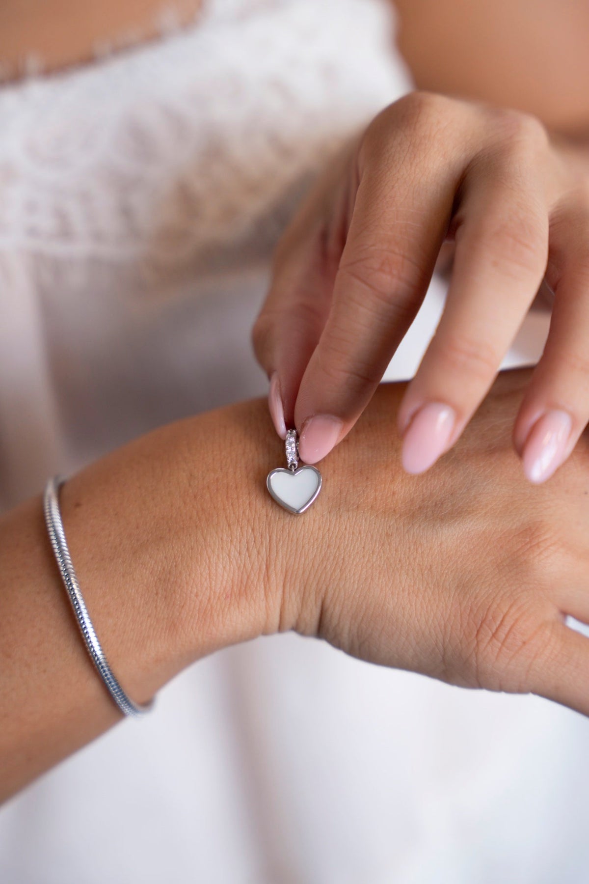 Fingers holding the silver Indy heart charm with breast milk, against a white lace top.
