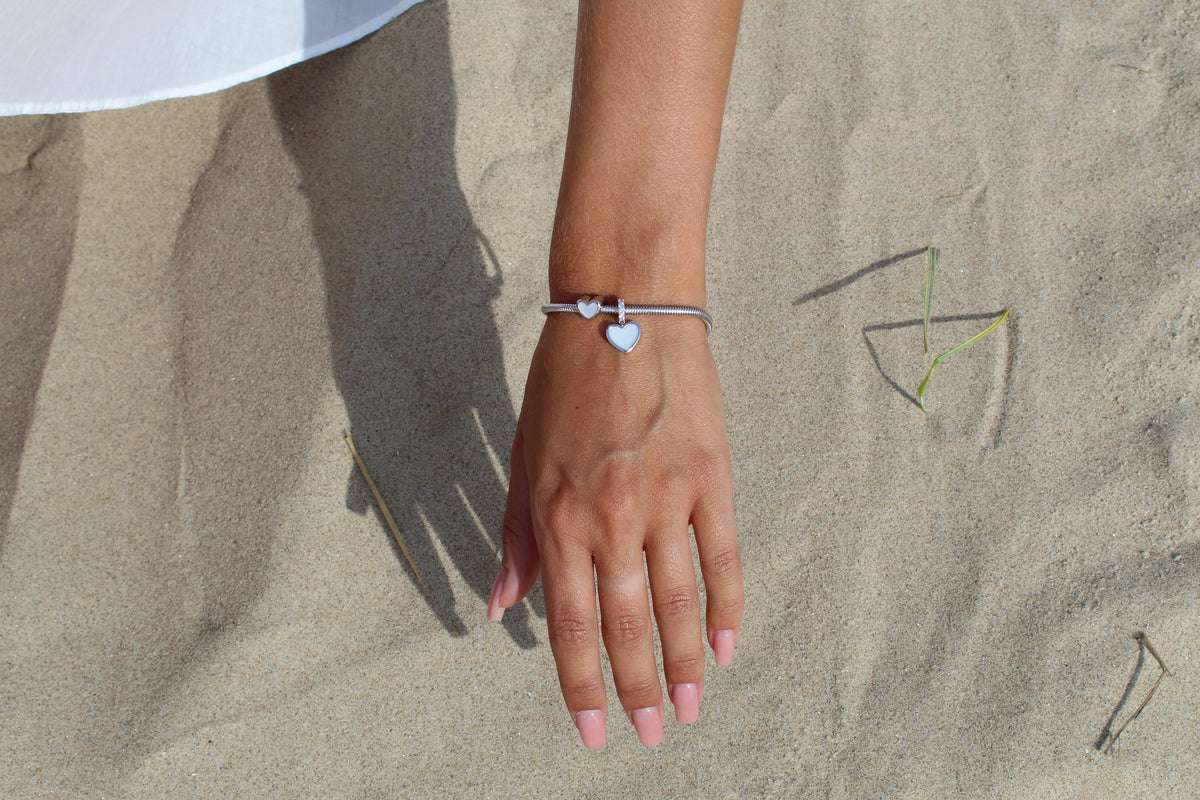Hand resting on sand, wearing a silver Indy heart breast milk charm on a silver snake chain bracelet