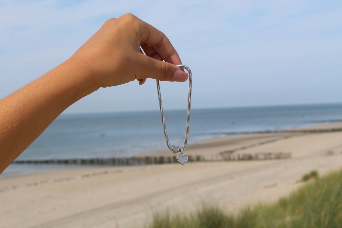 Silver bracelet with the Indy heart breast milk charm dangling from fingers above a beach
