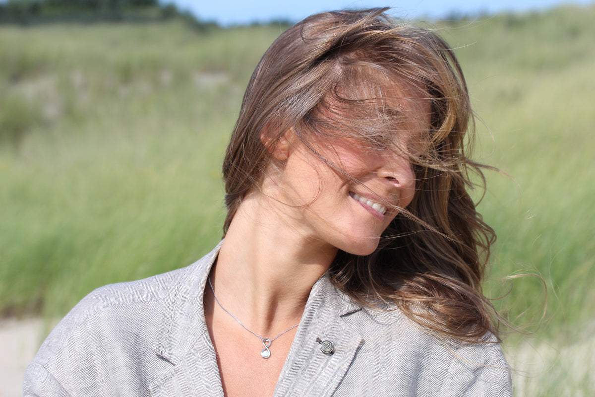 Smiling woman in the dunes with windswept hair wearing the silver Infinity breast milk pendant