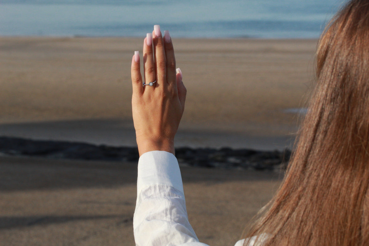 Raised hand facing the sea, wearing the silver Isabelle ring with a round milk stone