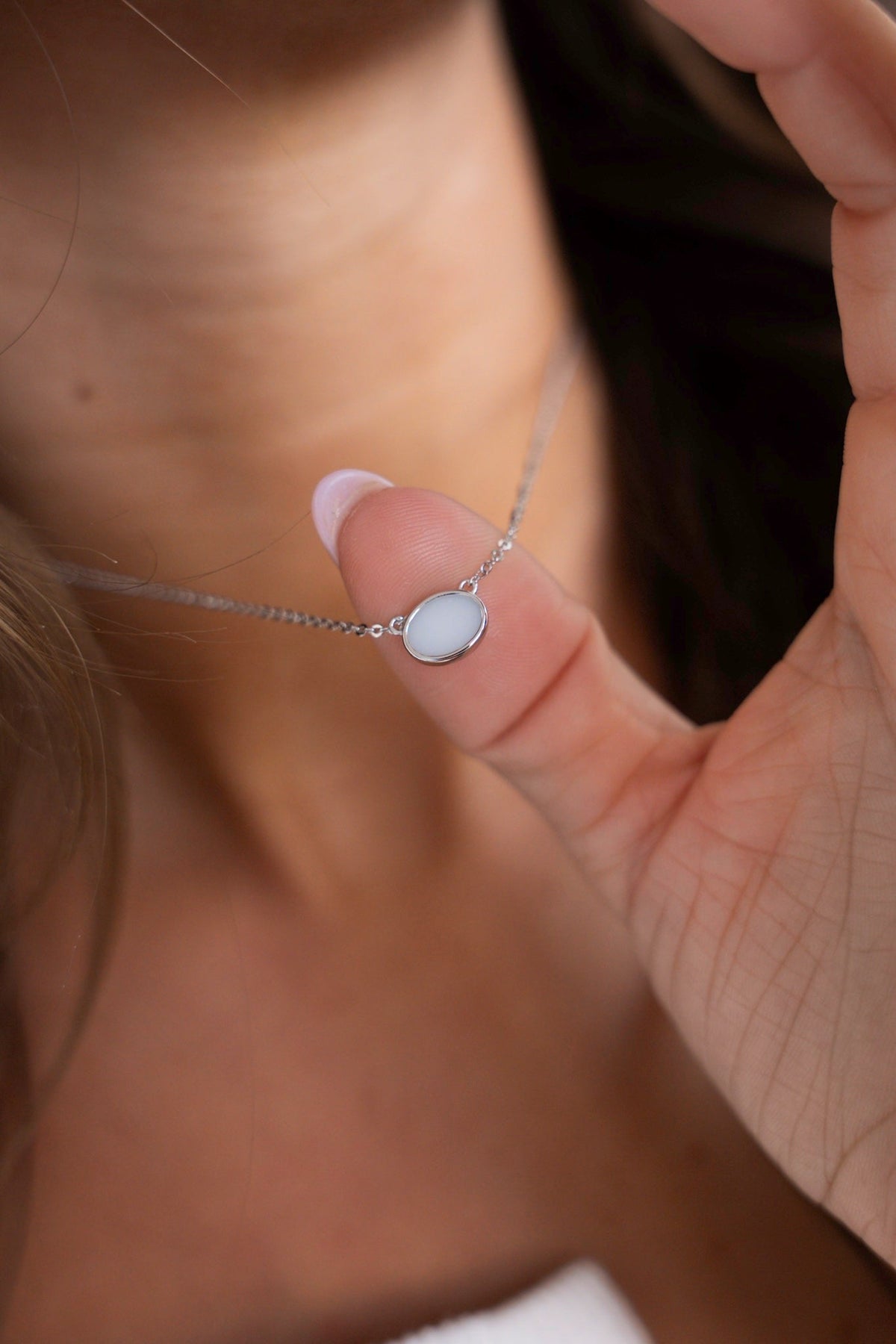 Fingers showing the Juna necklace in white gold with a sideways oval breast milk stone between two pieces of chain.