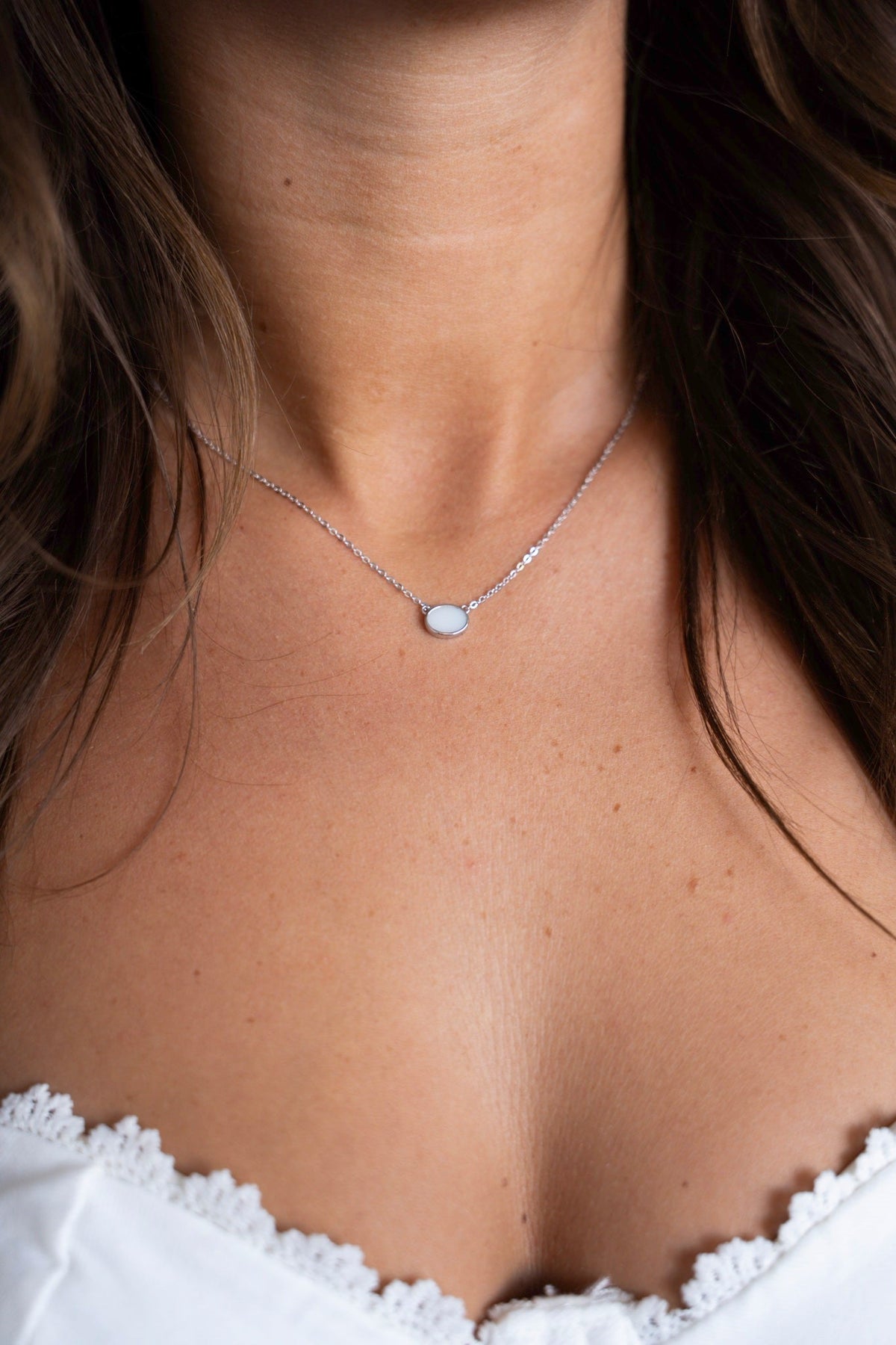 Juna necklace in white gold with a milky white oval stone on the collarbone, above a white lace top.