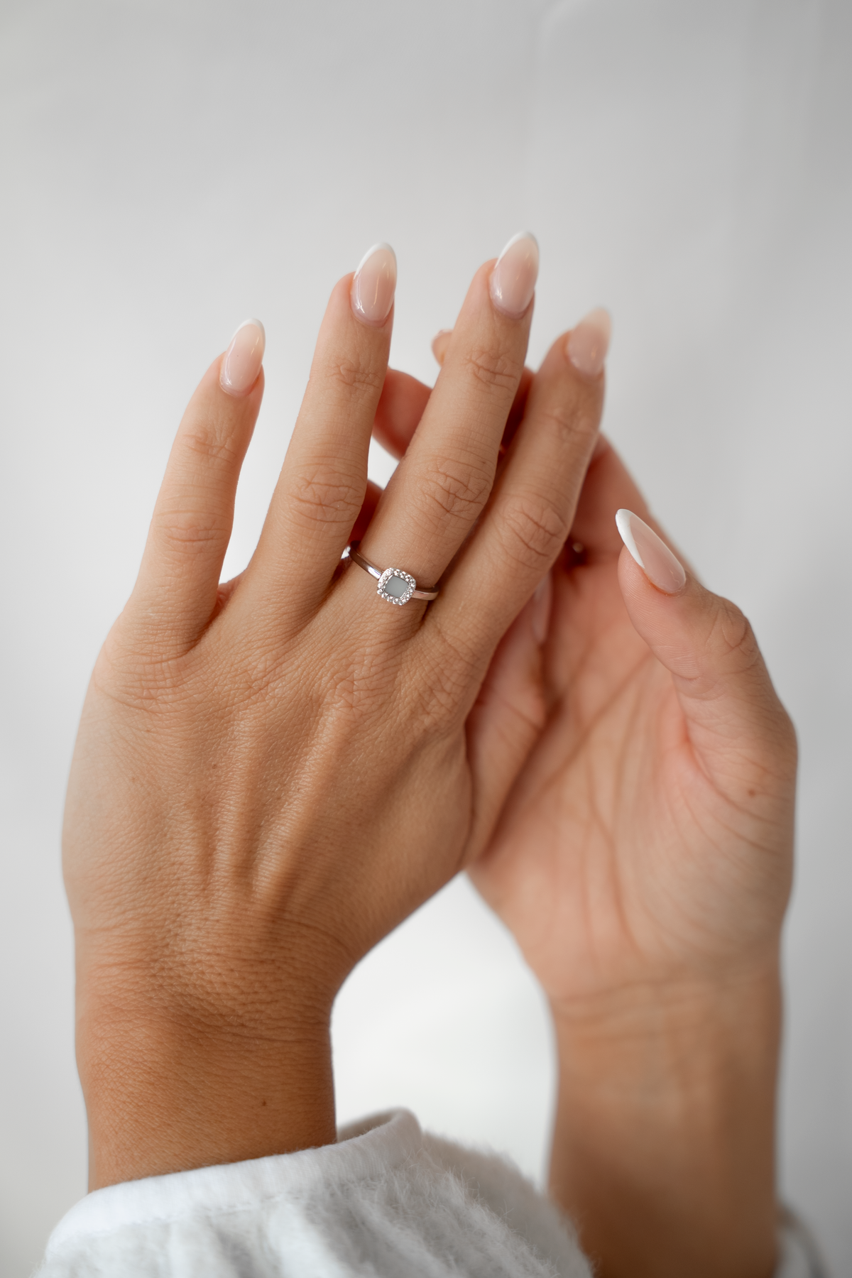 Two raised hands against a white wall, one wearing the silver Lara ring with a halo of clear stones