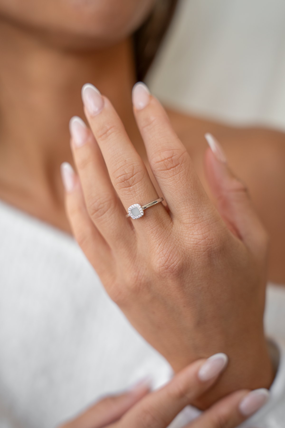 Hand held up to the chest wearing the silver Lara ring with a square milk stone and clear halo