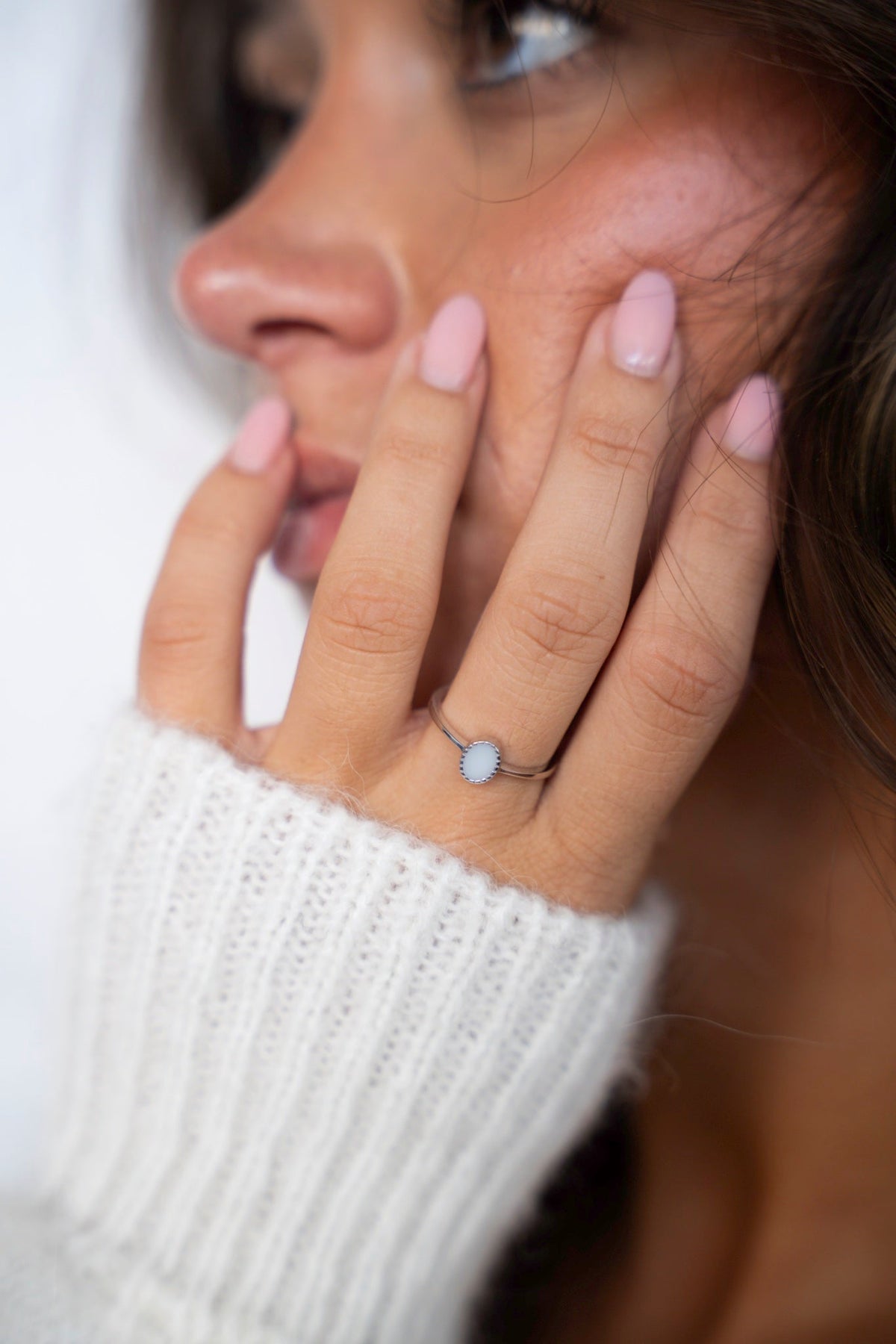 Lauren ring in silver with an oval breast milk stone in a fluted setting, hand resting on the cheek above a white knit sweater.