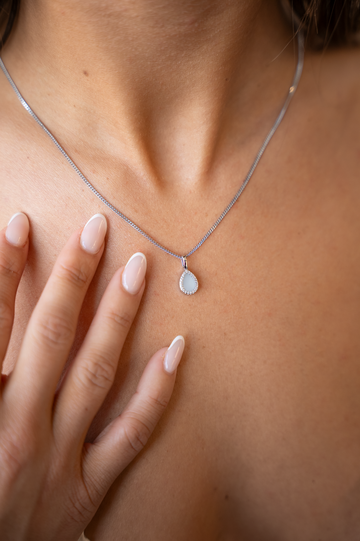 Close-up of the silver Lotte breast milk drop pendant on a fine chain, fingers resting below