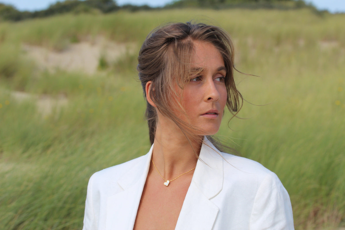 Woman looking sideways in the dunes, wearing a gold-colored Louisa heart breast milk necklace