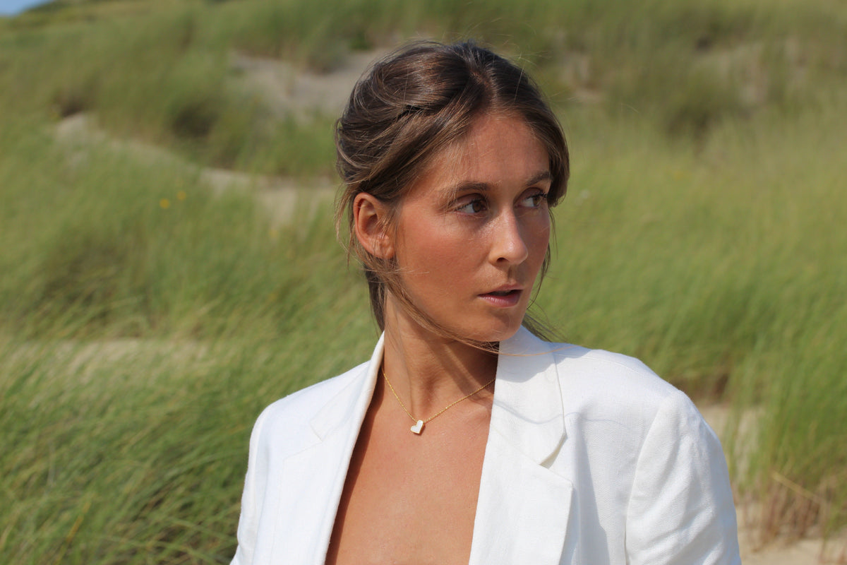 Woman in a white blazer among dune grass with a gold-colored Louisa tilted heart necklace