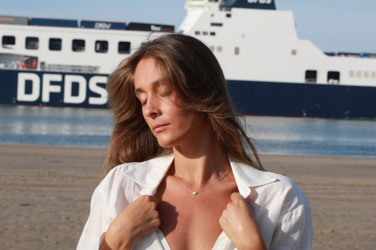 Woman with closed eyes in front of a ship on the shore, wearing a Luna oval breast milk necklace