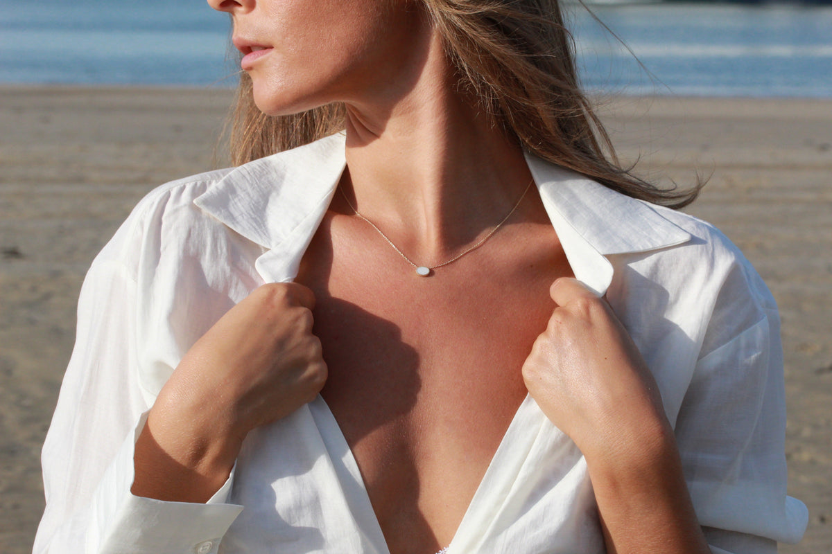 Luna necklace with an oval breast milk stone below the collarbone, white shirt and beach behind