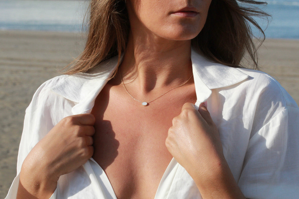 Woman on the beach holding her white shirt collar, Luna oval breast milk necklace on her chest