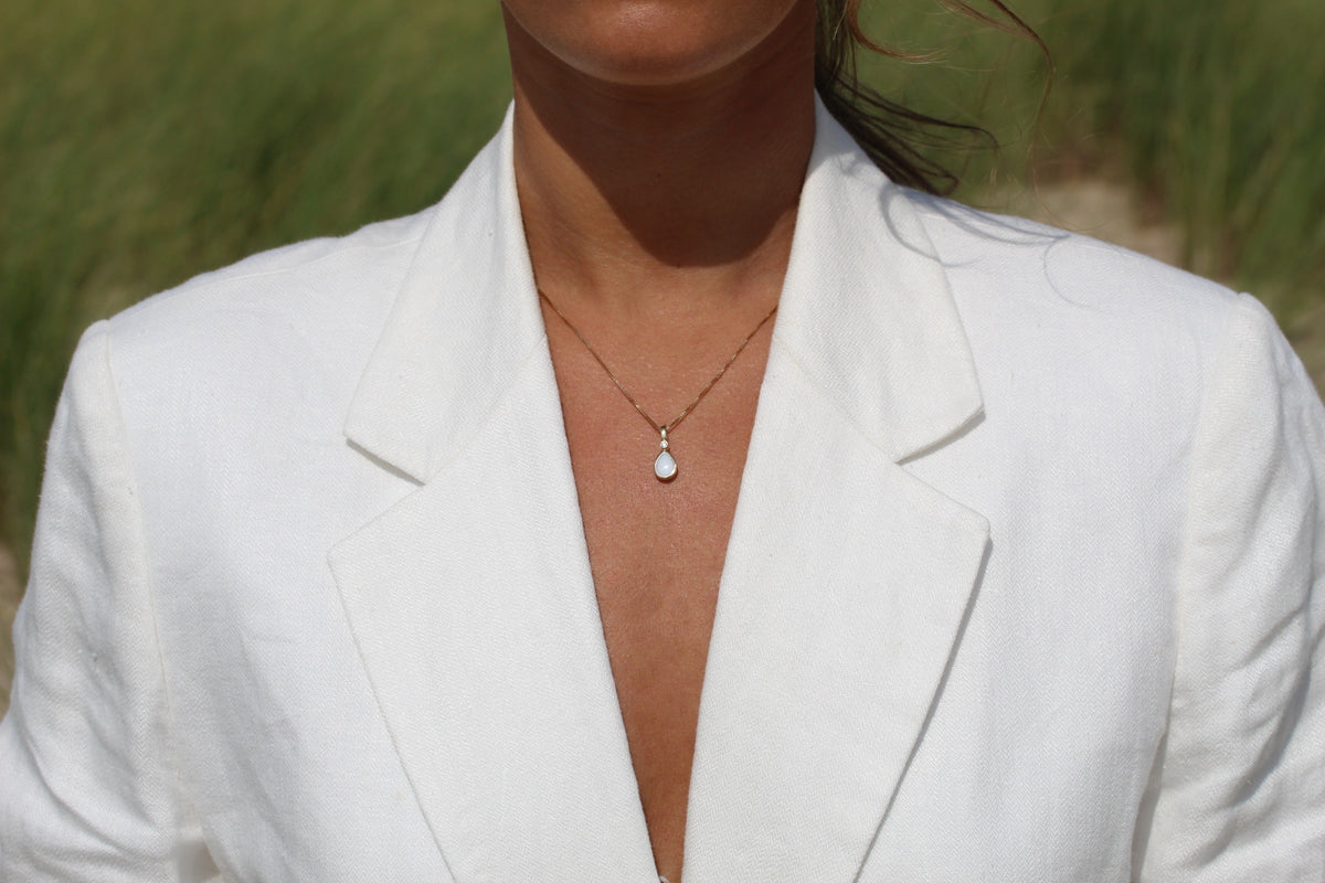 Magic Drop gold teardrop breast milk pendant on a fine chain, worn with an open white blazer in the dunes