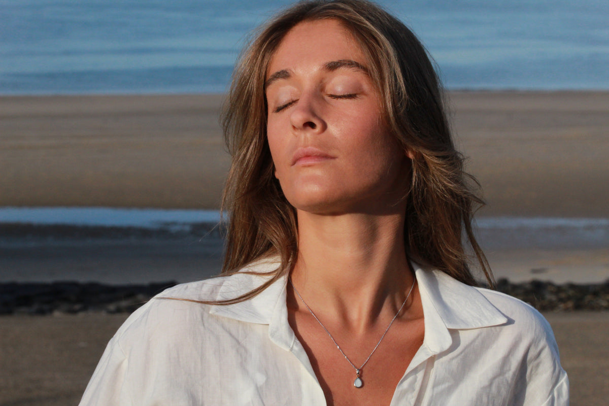 Woman with closed eyes on the beach wearing a silver-colored Magic Drop teardrop breast milk pendant