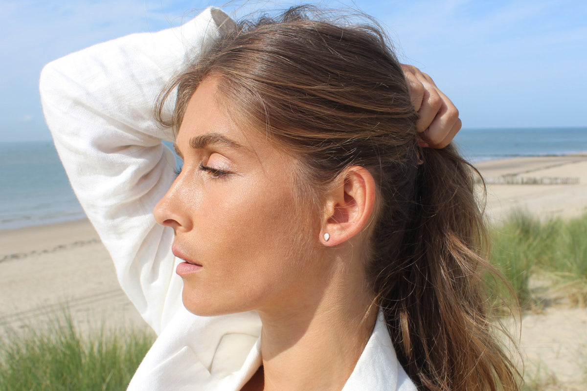 Woman on a beach with her arm raised, wearing a small Mini Drop breast milk stud earring