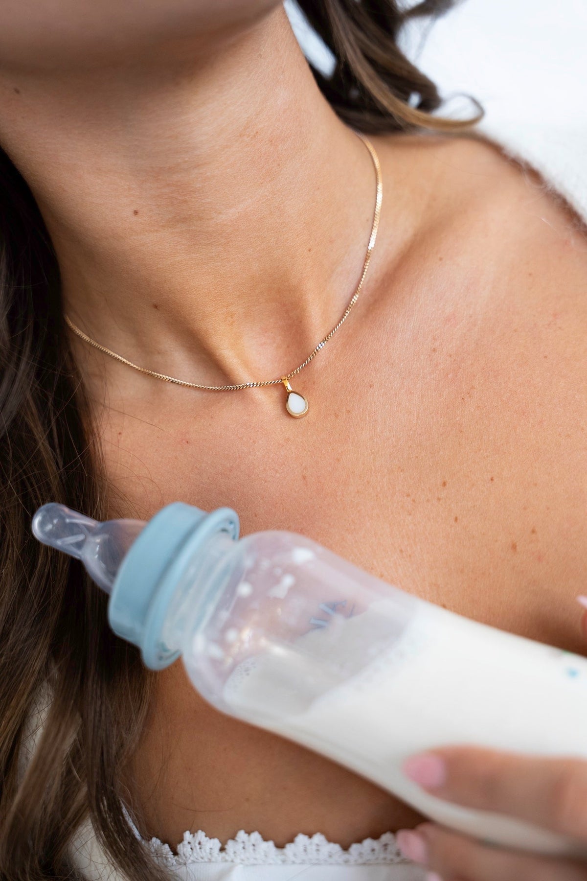 Gold Pure Drop pendant with a teardrop-shaped breast milk stone at the neck, next to a baby bottle of milk.