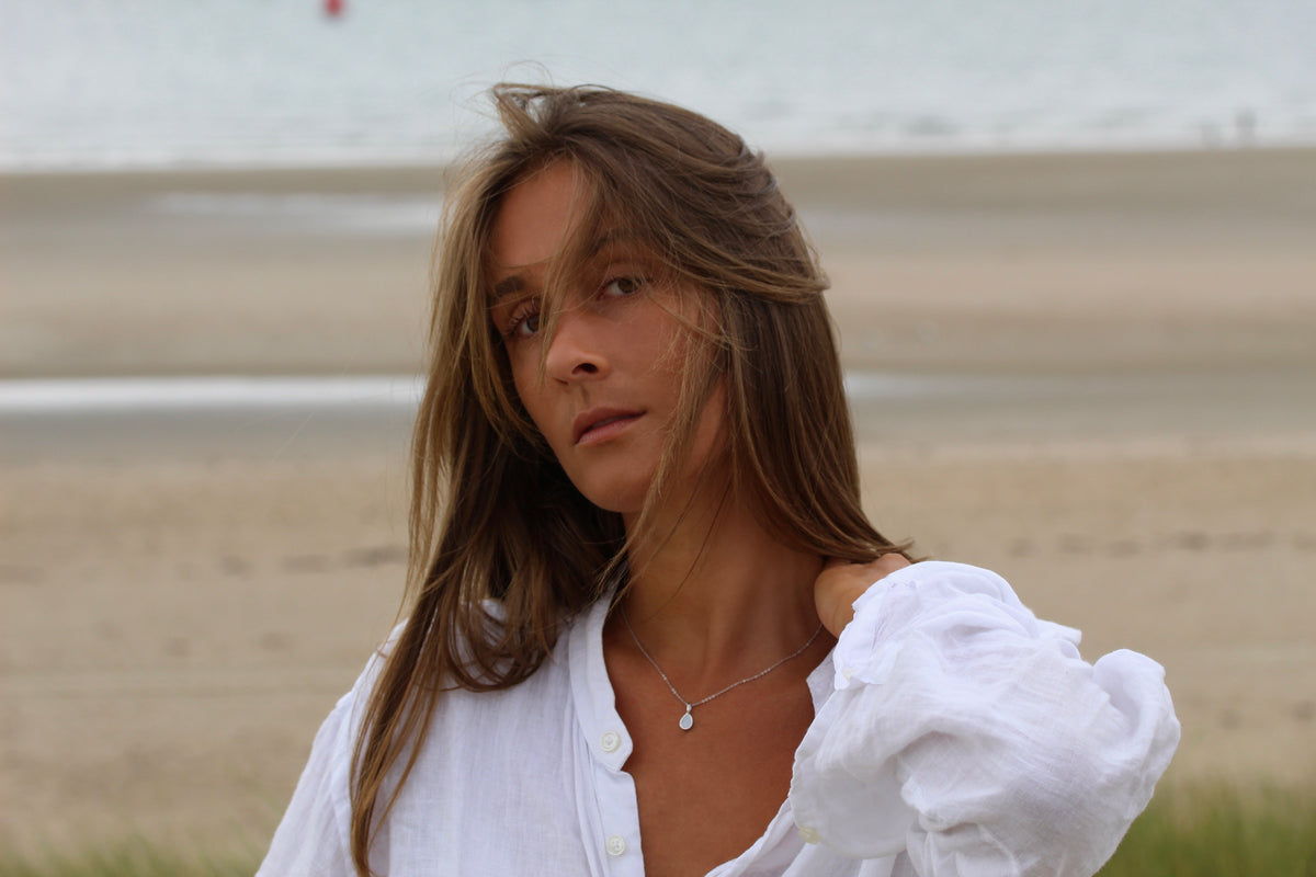 Woman on the beach in a white linen blouse wearing the silver-tone Pure Drop breast milk pendant