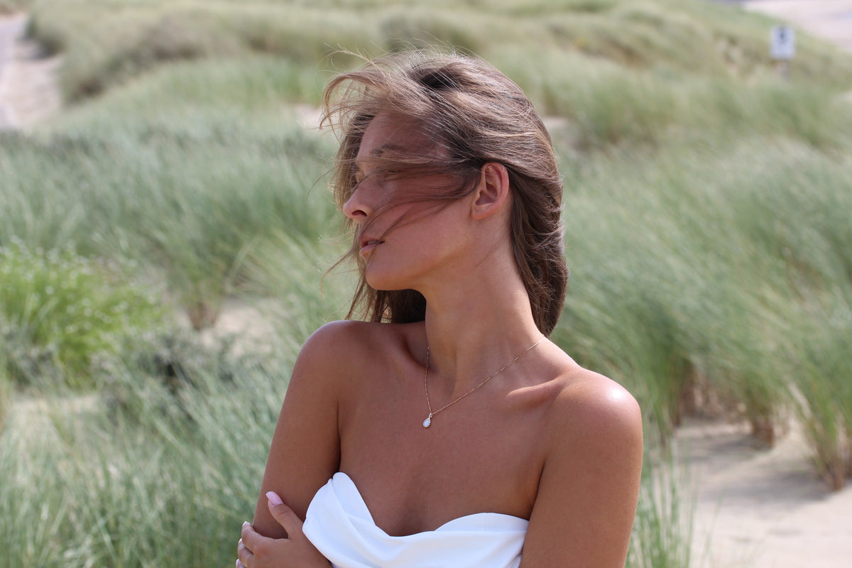 Woman in a strapless white top in the dunes wearing the Pure Drop breast milk pendant on a fine chain