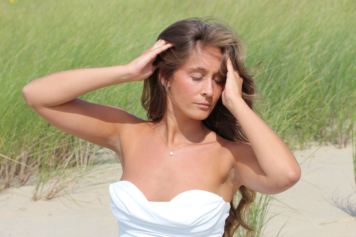 Woman with hands in her hair in the dunes wearing the Pure Drop breast milk pendant in gold