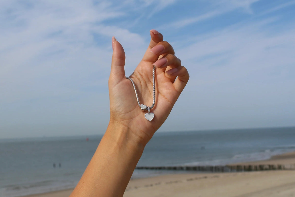 Silver charm bracelet featuring two heart-shaped charms, held in a hand against a beach and ocean backdrop.