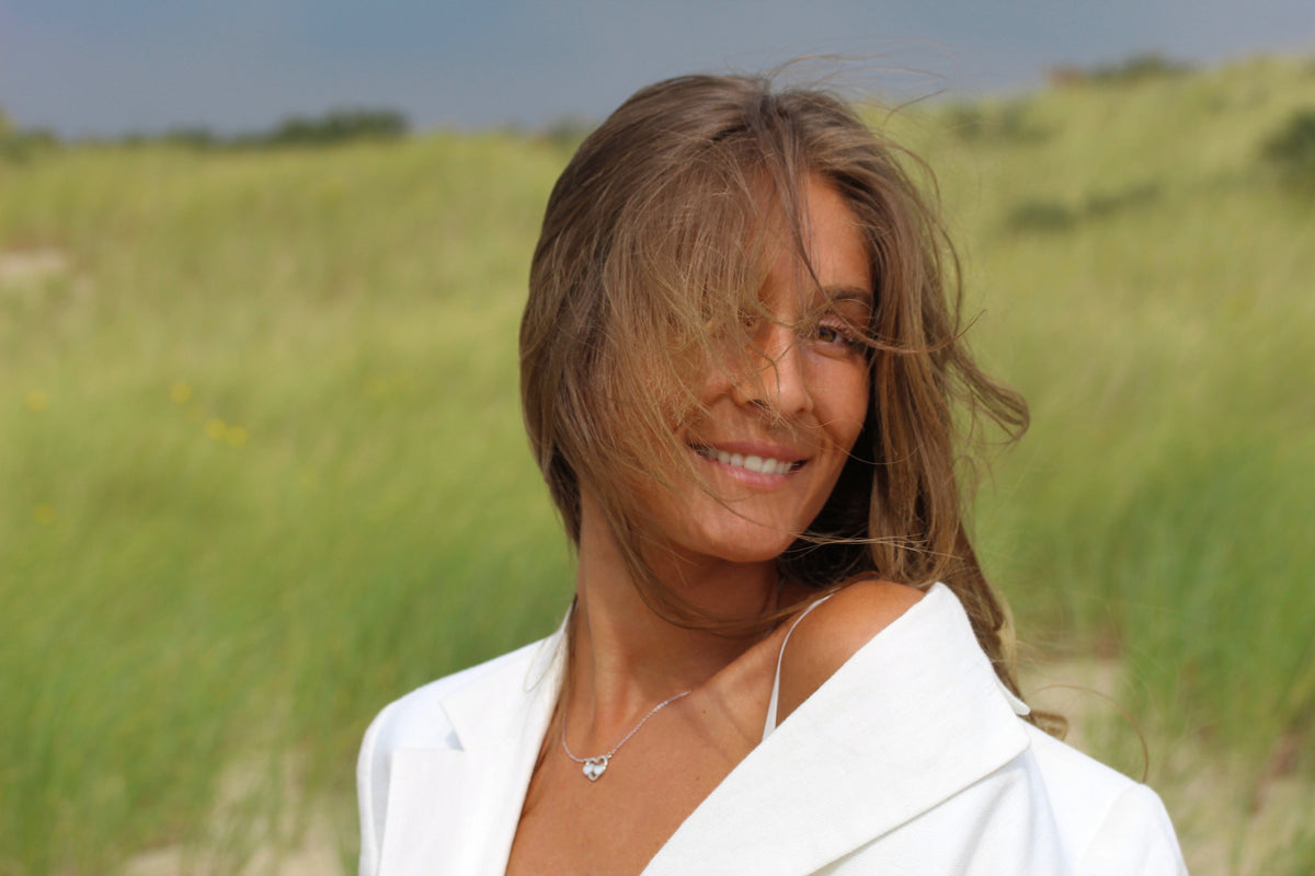 Smiling woman in the dunes wearing a silver-colored Trinity open heart breast milk necklace