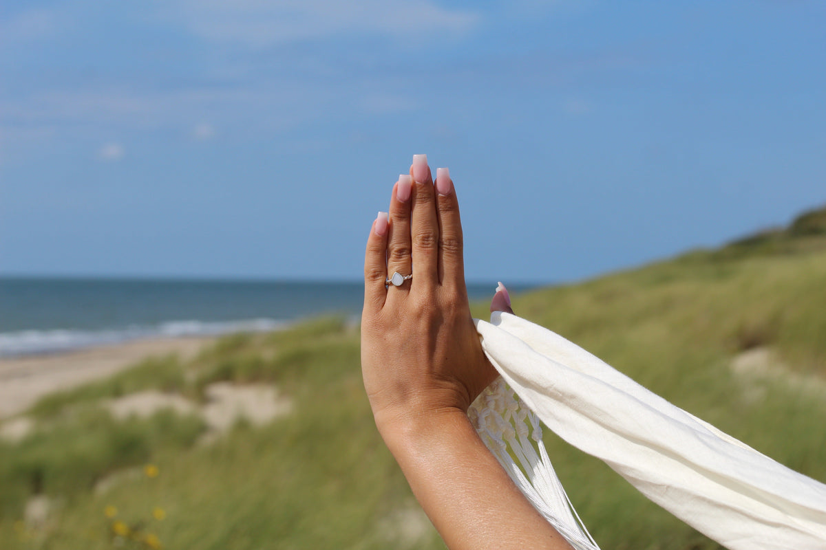 Silver Turning Droplet breast milk ring on the finger, raised hand against blue sky, dunes and sea