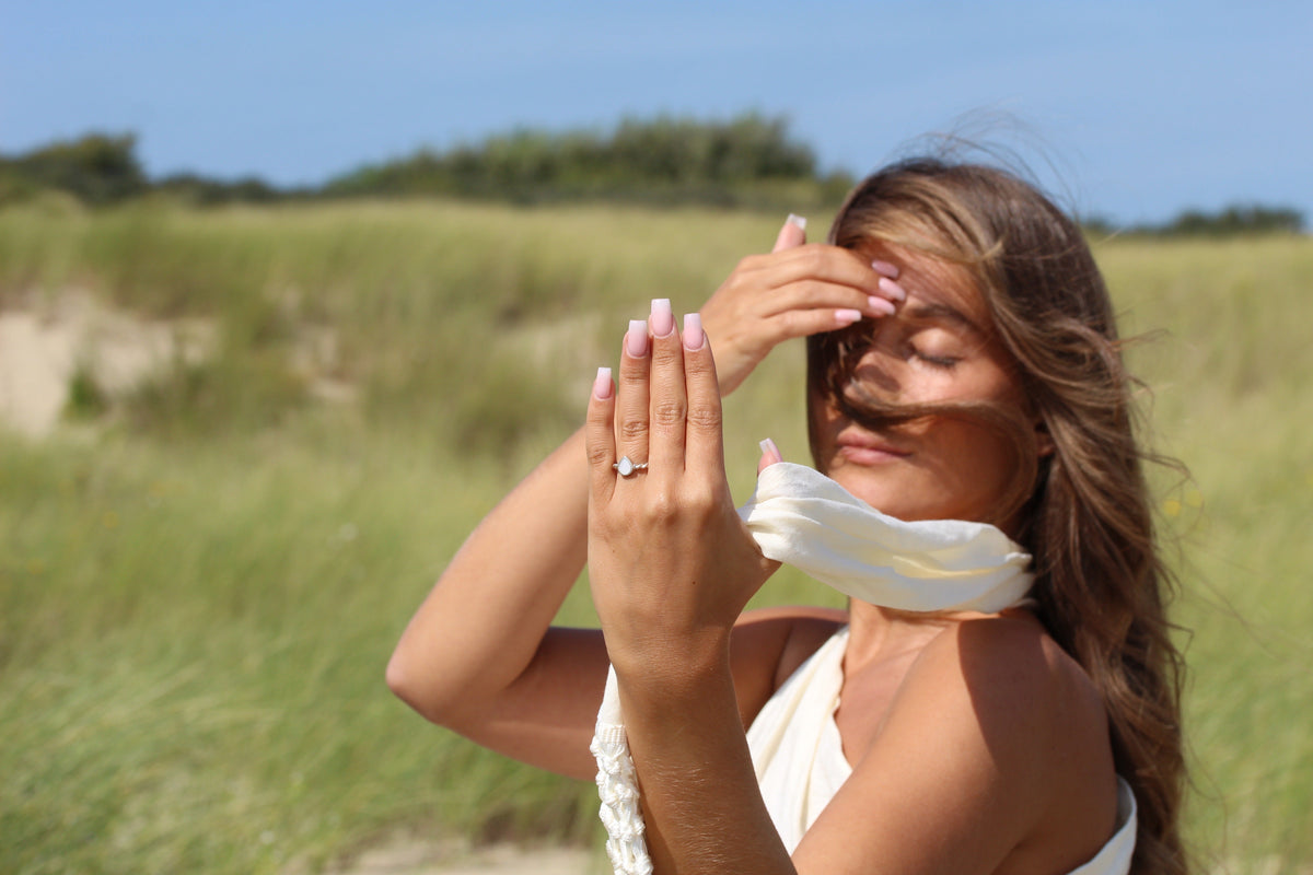 Silver Turning Droplet breast milk ring on the finger, raised hand with a cream scarf in the dunes
