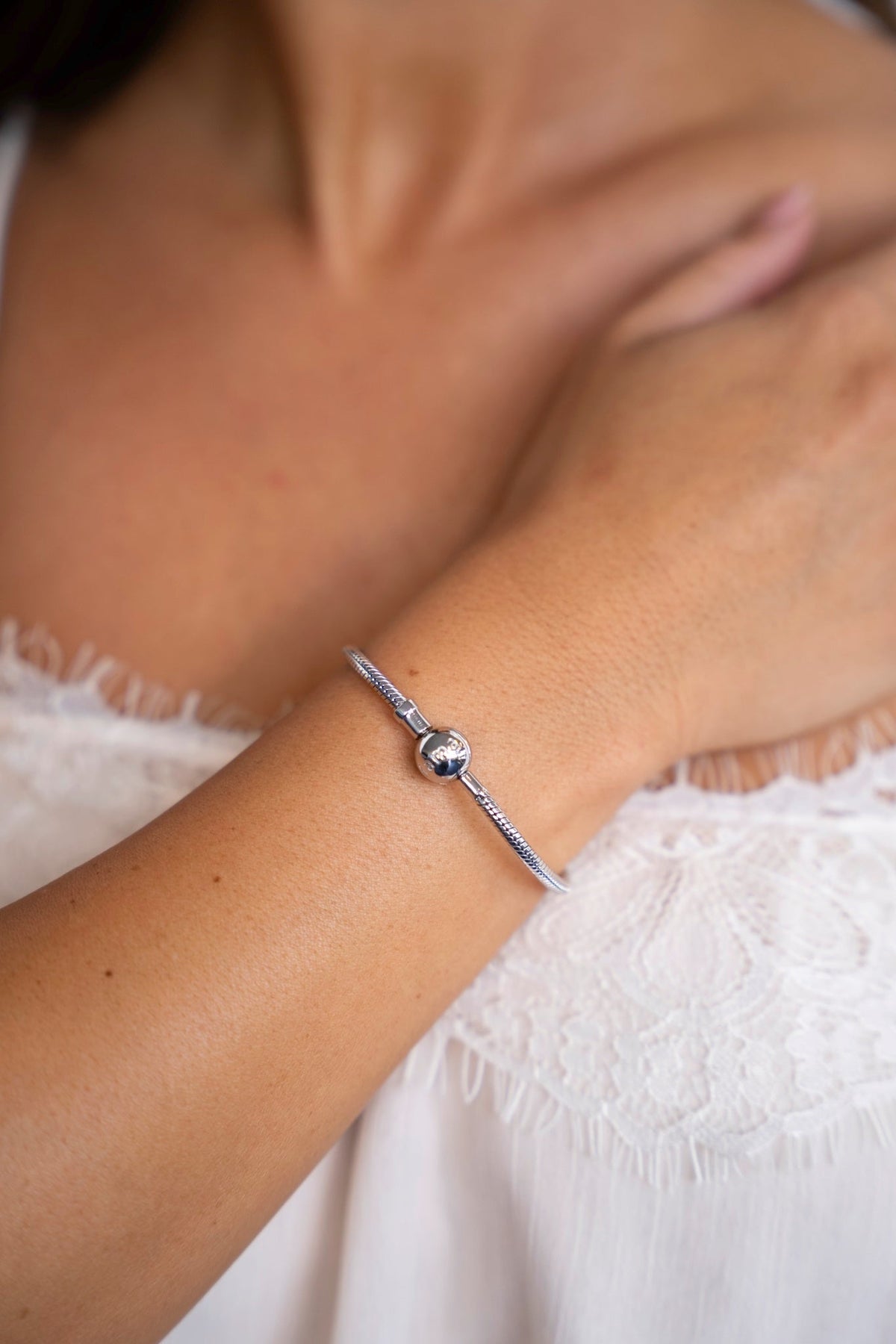Silver charm bracelet on the wrist, the hand resting on the chest above a white lace top.